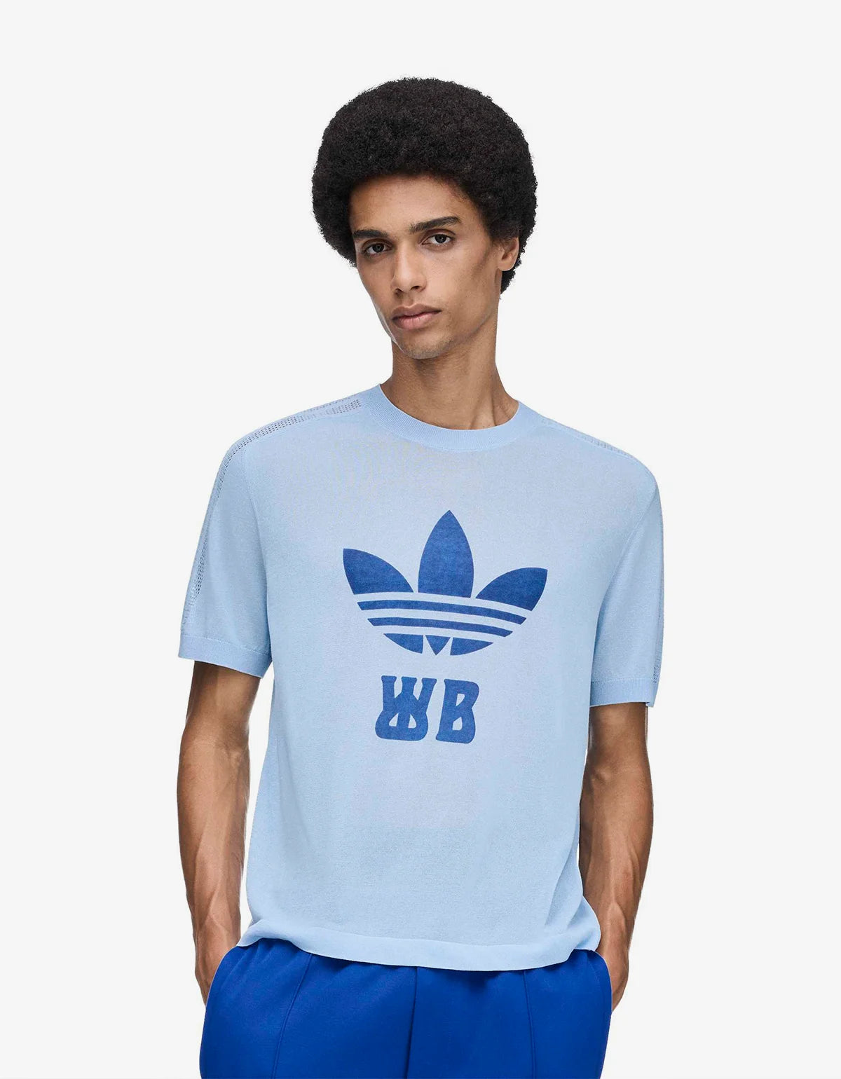 A man wearing a light blue crewneck t-shirt with a blue Adidas Trefoil logo and 'WB' monogram on the front, paired with royal blue pants, against a light gray studio background.
