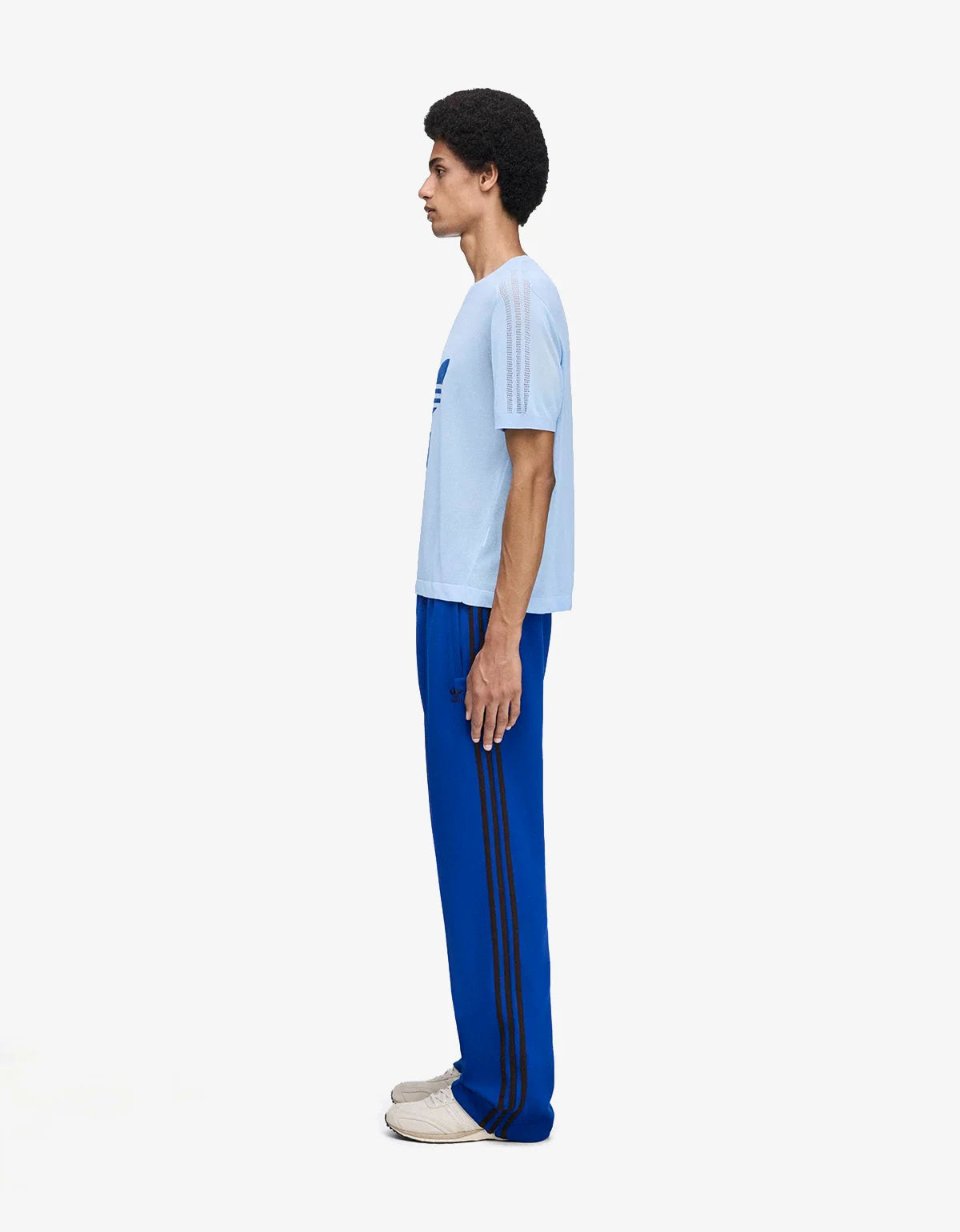 A model shown in profile wearing a light blue t-shirt and bright blue track pants with black stripes, standing against a plain white background.