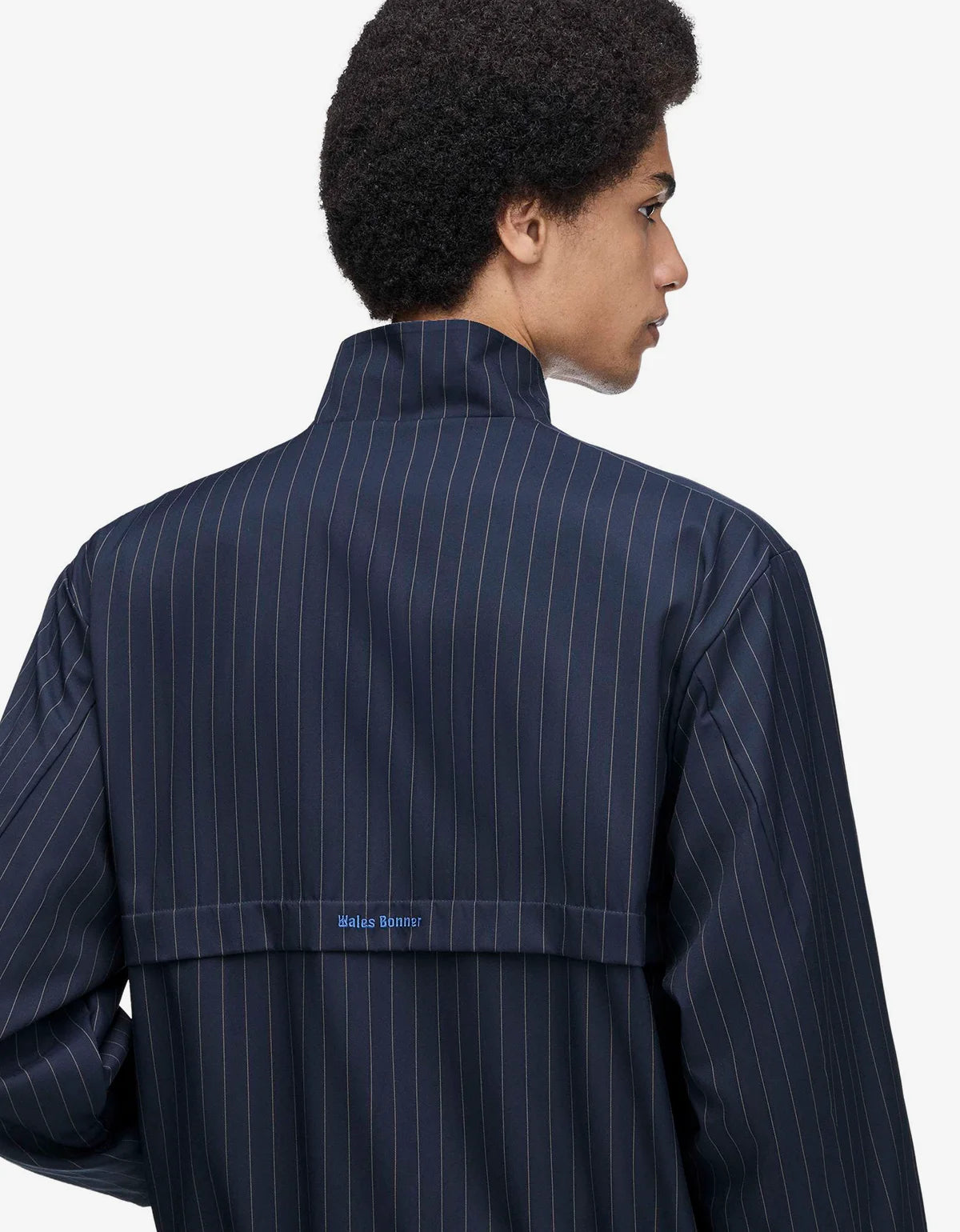 A man shown from the back wearing a navy blue pinstriped jacket with a high collar and a small blue 'Wales Bonner' logo on the back waistband, against a plain light gray background.