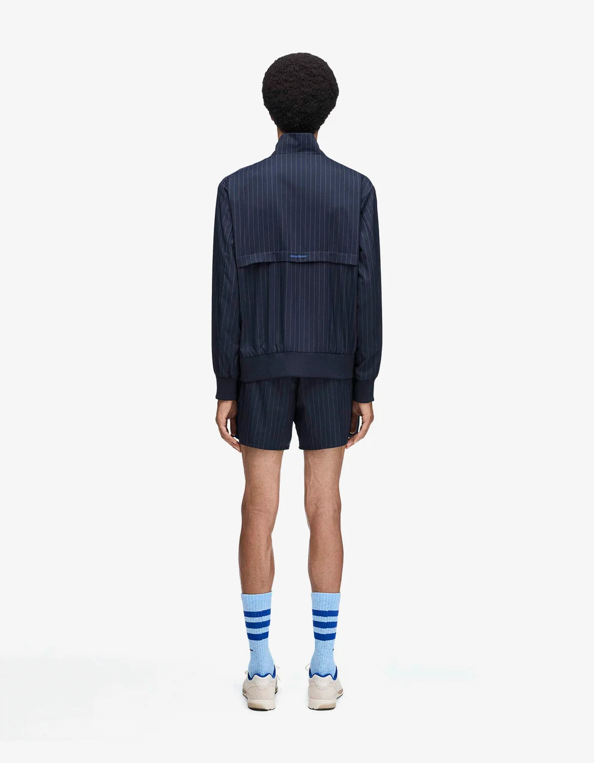 A model shown from the back wearing a dark navy blue pleated zip-up jacket and matching pleated shorts, paired with light blue and white striped socks and white sneakers, against a plain white background.