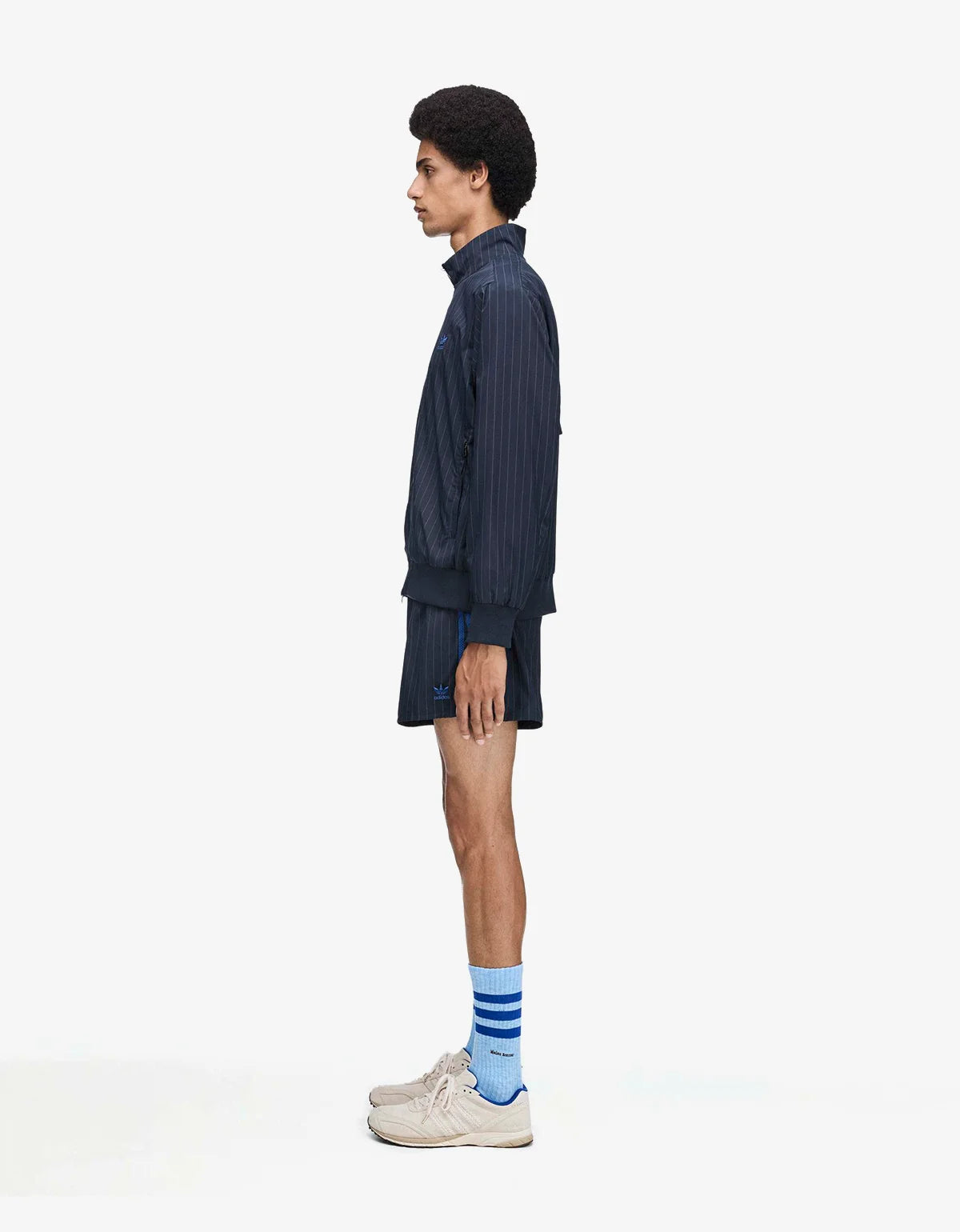 A model shown in profile wearing a dark navy blue pinstriped zip-up jacket, matching shorts, blue and white striped socks, and beige sneakers against a plain white studio background.