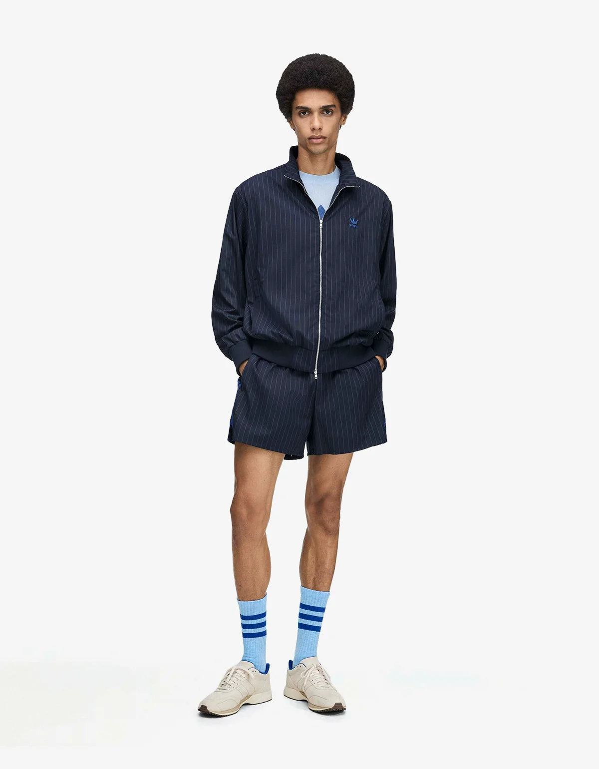 A model stands against a light gray studio background wearing a matching navy pinstriped zip-up jacket and shorts set, a light blue t-shirt, blue striped socks, and beige sneakers.