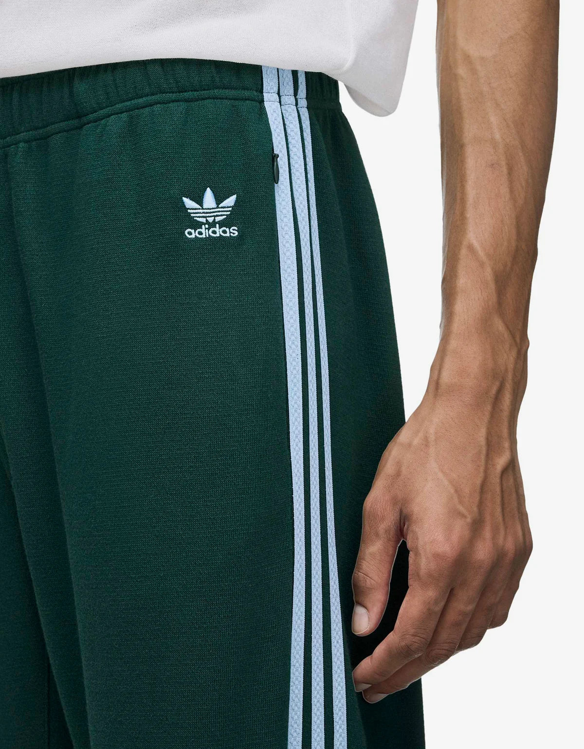 Close-up of a person wearing dark green Adidas track pants with white side stripes and a white Adidas logo patch on the thigh, paired with a white top against a light gray background.