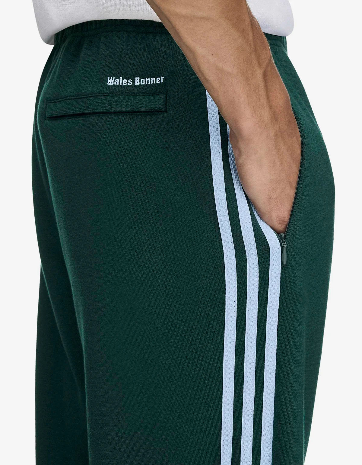 Close-up of a person wearing dark green track pants with three light blue stripes down the side, with the text 'Wales Bonner' printed on the back pocket.