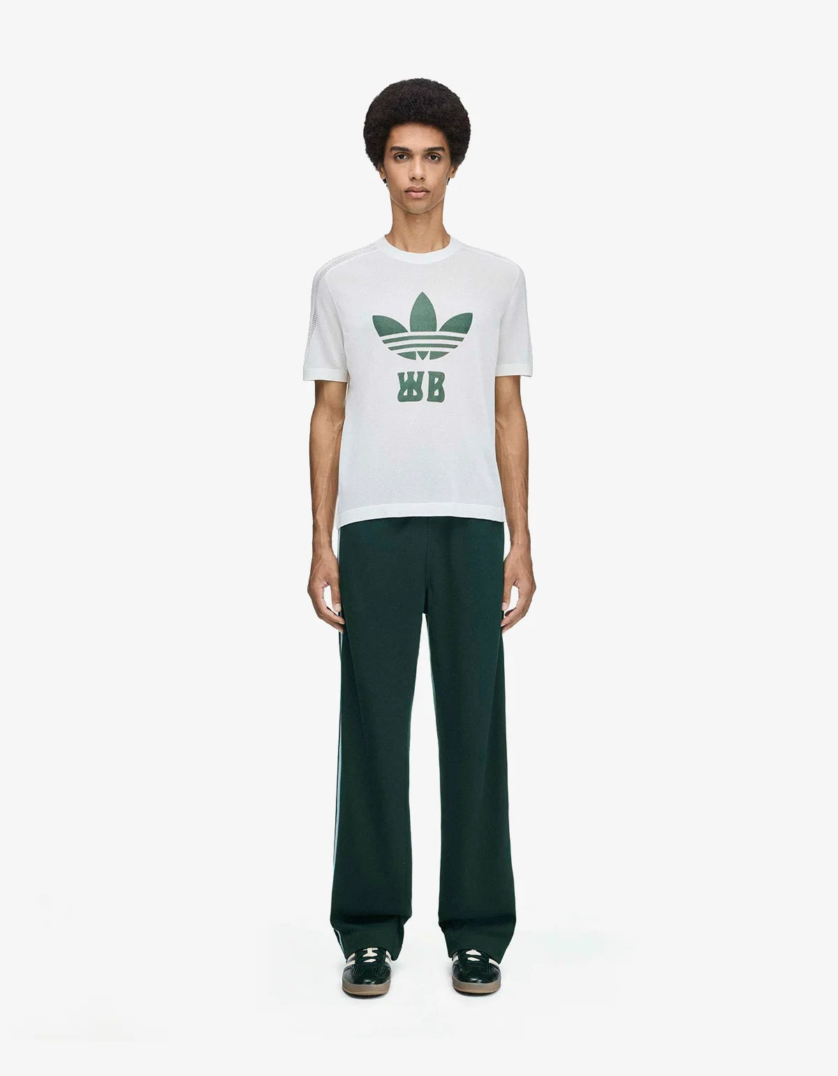 A model stands against a light gray studio background wearing a white short-sleeve t-shirt with a green trefoil logo and 'WB' text on the chest, paired with dark green wide-leg pants and black sneakers.