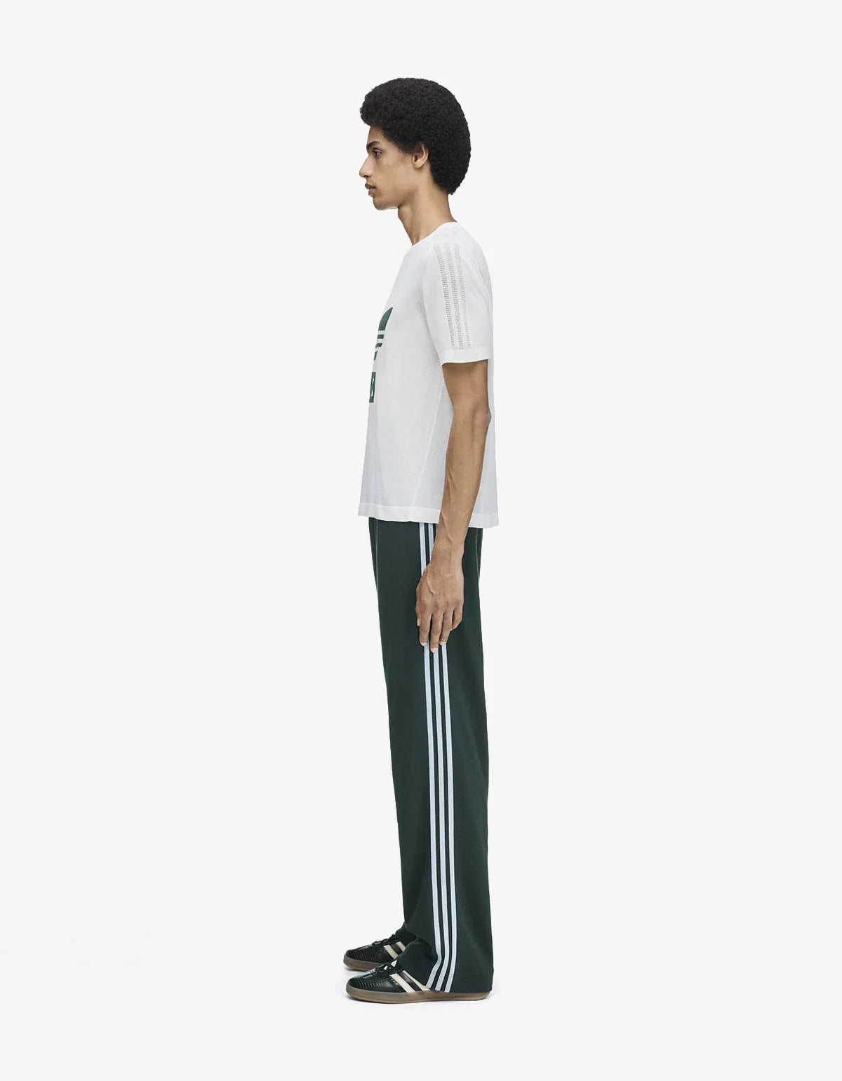 A man in profile wearing a white t-shirt and dark green track pants with blue side stripes, standing against a plain light gray background.