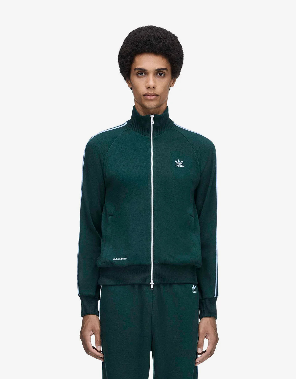 A model wearing a dark green Adidas track jacket with blue stripes, standing against a plain white studio background.