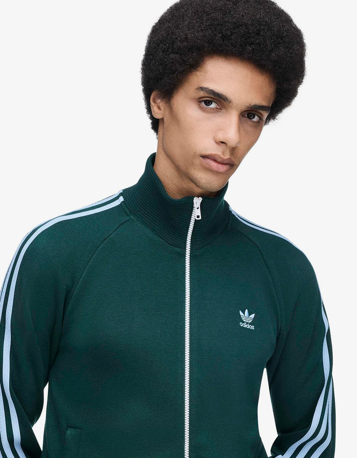A model wearing a dark green Adidas track jacket with blue stripes, standing against a plain white studio background.
