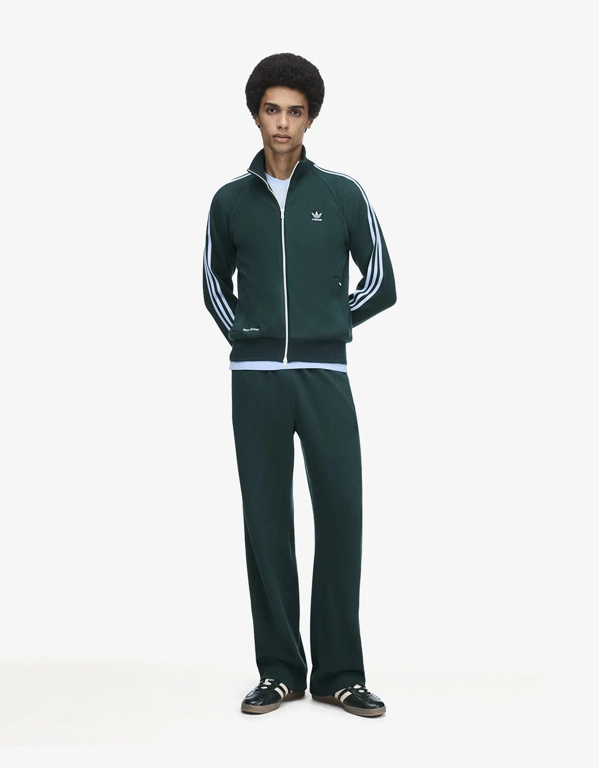 A model wearing a dark green Adidas track jacket and matching track pants with light blue stripes, standing against a plain white studio background.