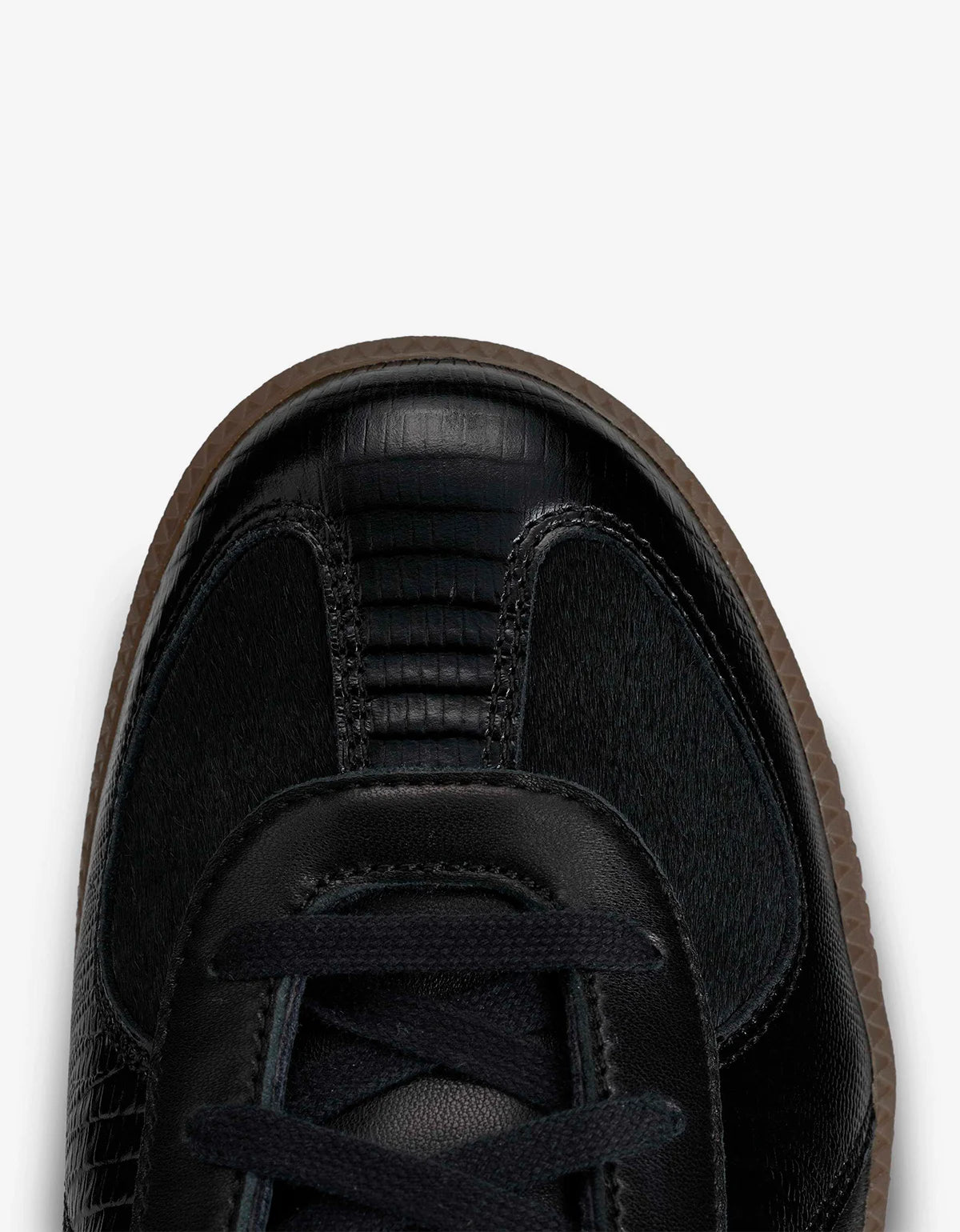 Close-up top view of a black sneaker showing the toe area, textured leather panels, black laces, and a brown rubber sole against a light gray background.