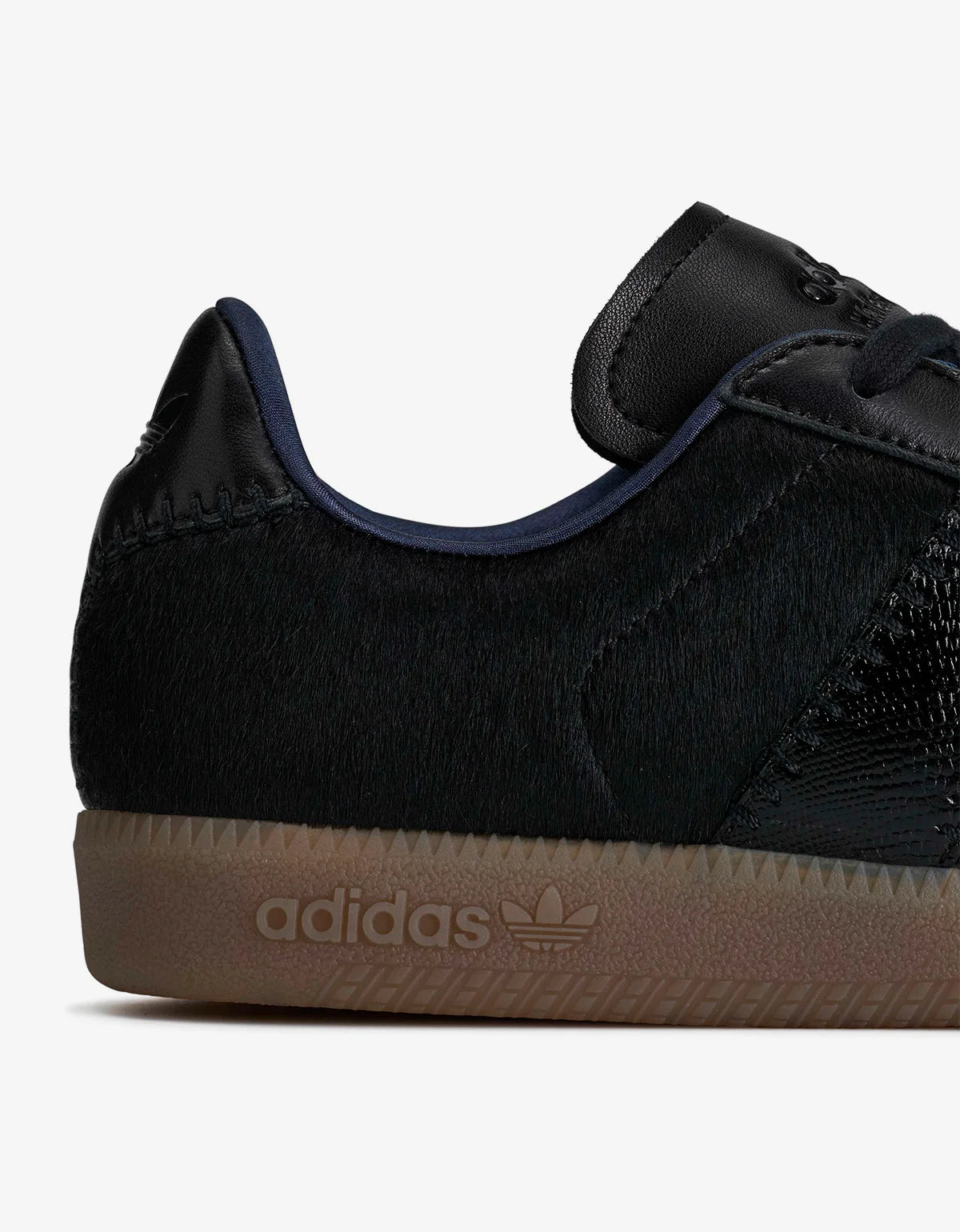 Close-up side view of an Adidas sneaker showing the heel, black textured upper, navy blue collar lining, and a brown gum sole with embossed adidas branding.