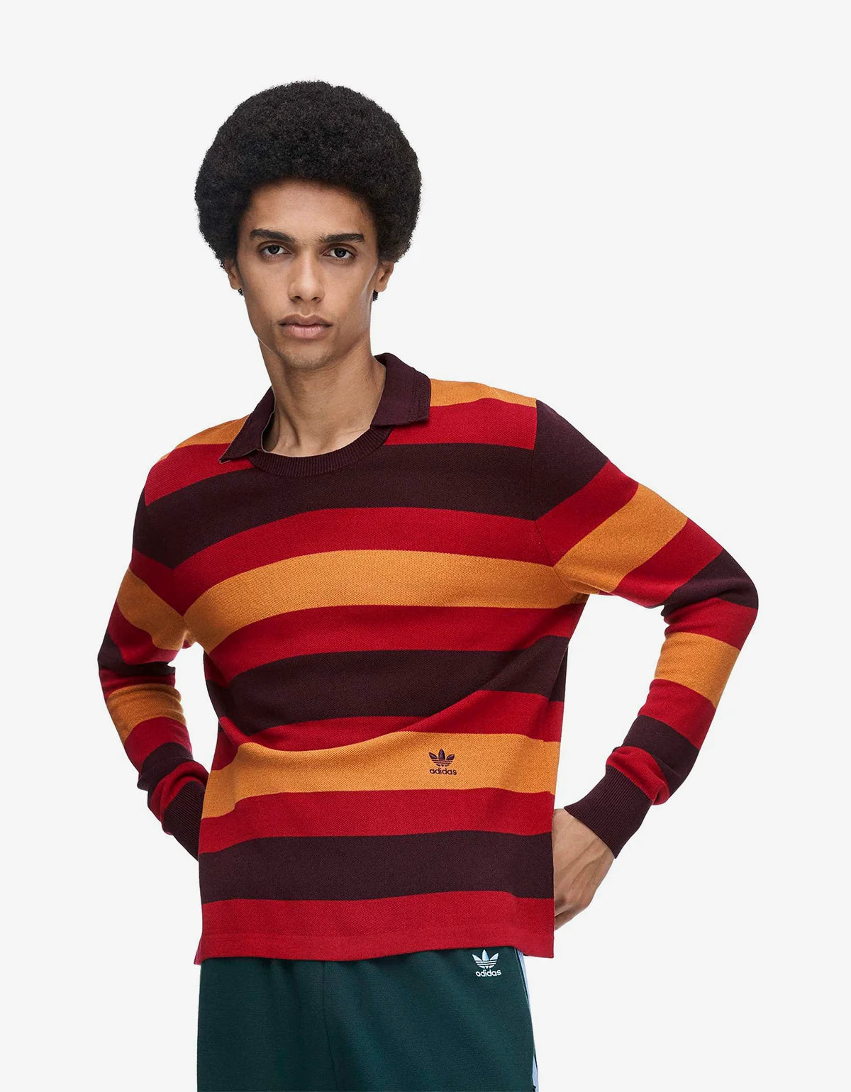 A model wearing a long-sleeve striped knit sweater with horizontal bands of dark red, orange, and dark maroon, featuring a small black Adidas trefoil logo on the left chest.