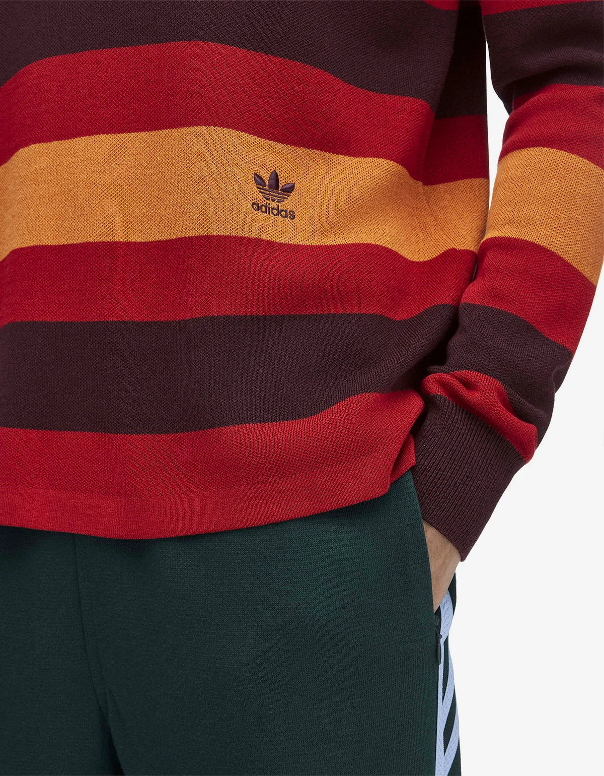 Close-up of a person wearing a striped knit sweater with horizontal bands of dark brown, red, and mustard yellow, featuring a small black Adidas trefoil logo on the chest. The person is also wearing dark green pants with white side stripes.