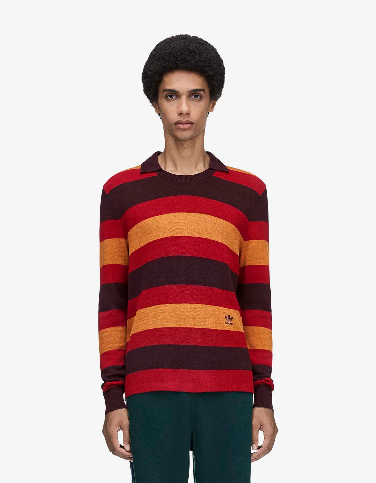 A model wearing a long-sleeve striped knit sweater with horizontal bands of red, orange, and dark burgundy, featuring a small embroidered Adidas trefoil logo on the lower right chest.