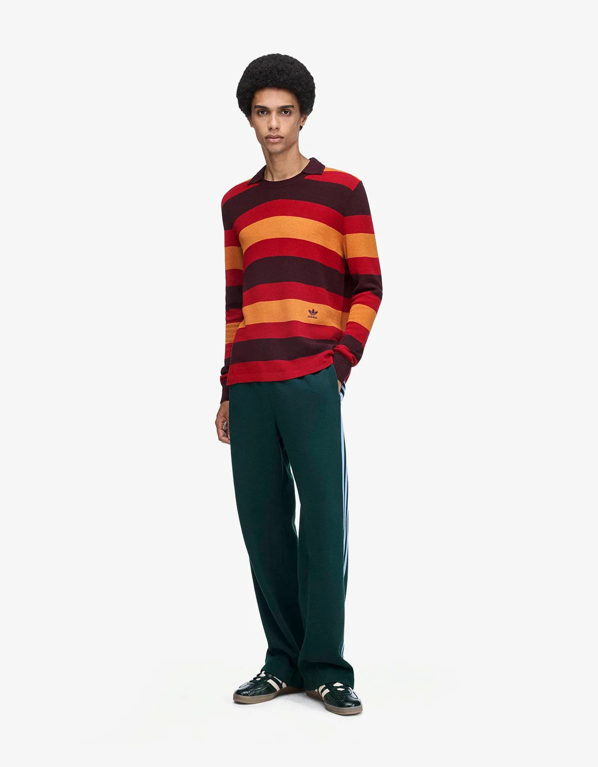A model stands against a light gray studio background wearing a long-sleeve crewneck sweater with bold horizontal stripes in red, orange, and dark brown, paired with dark green track pants and dark sneakers.