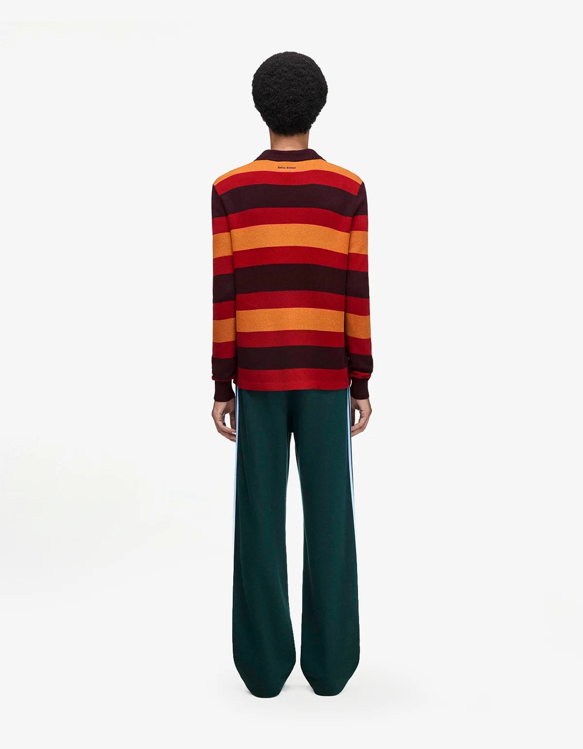 A model shown from the back wearing a long-sleeve knit sweater with horizontal stripes in red, orange, and dark brown, paired with dark green wide-leg trousers, against a plain white studio background.