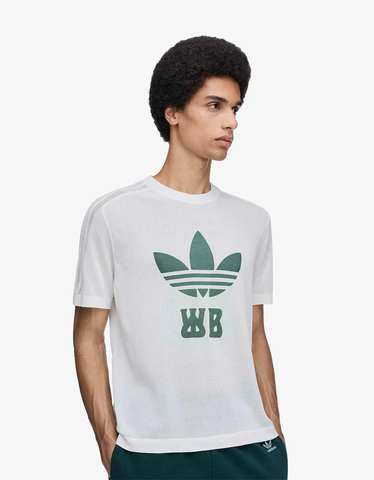 A man wearing a white short-sleeve t-shirt with a green Adidas Trefoil logo and 'WB' monogram on the front, paired with dark green pants, against a light gray studio background.