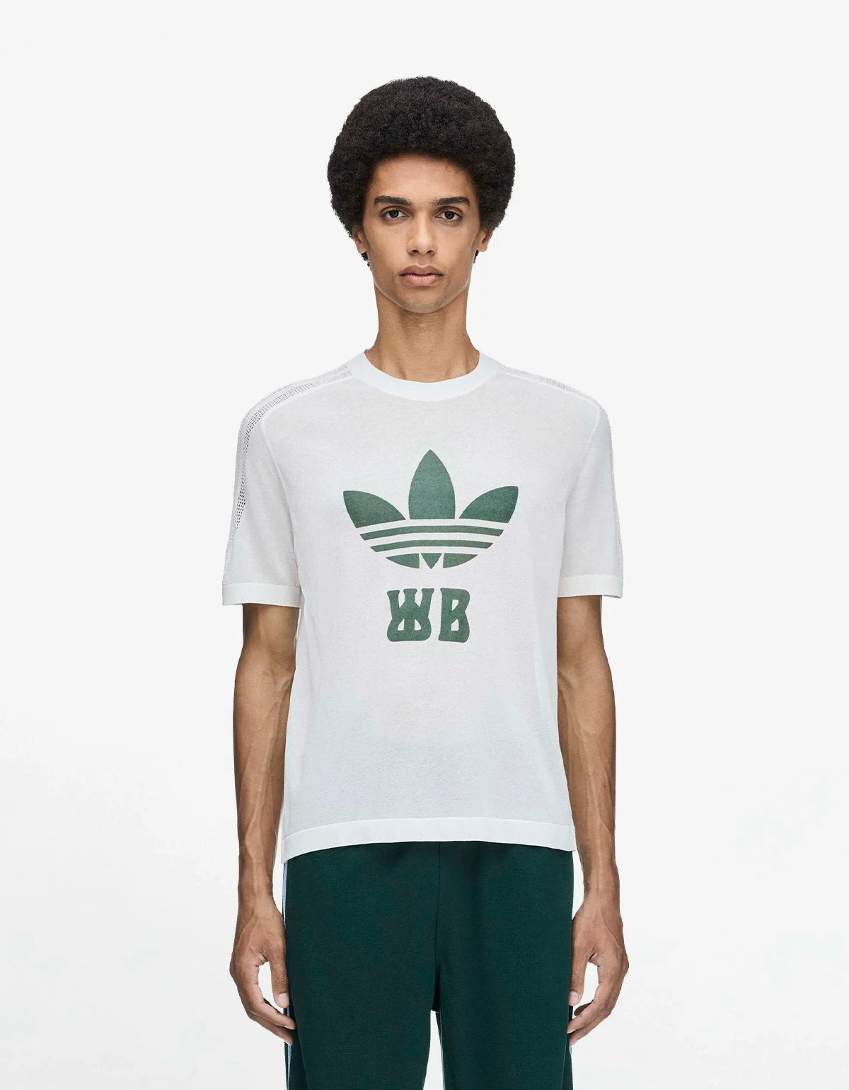 A man wearing a white short-sleeve t-shirt with a green Adidas Trefoil logo and 'WB' monogram on the front, paired with dark green pants, against a light gray studio background.