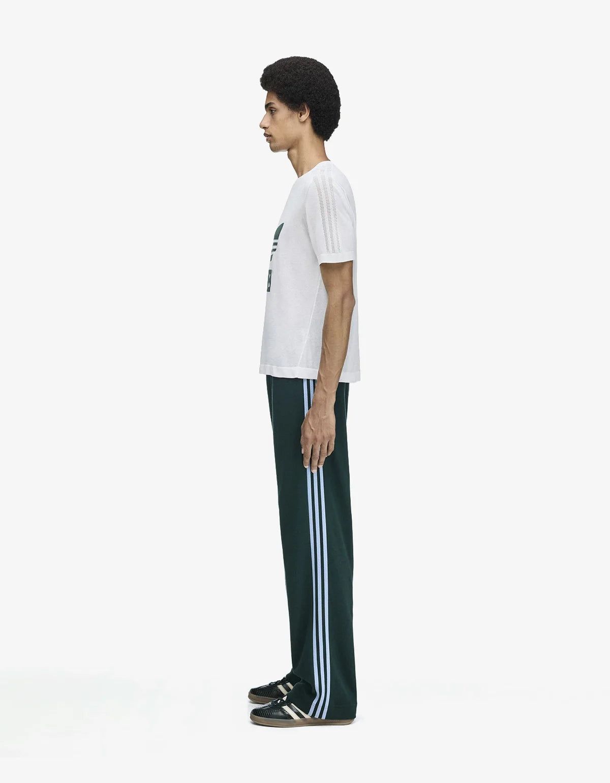 A model shown in profile wearing a white short-sleeve t-shirt with a green graphic on the chest and dark green track pants featuring three white side stripes, paired with black and white sneakers, against a plain white studio background.