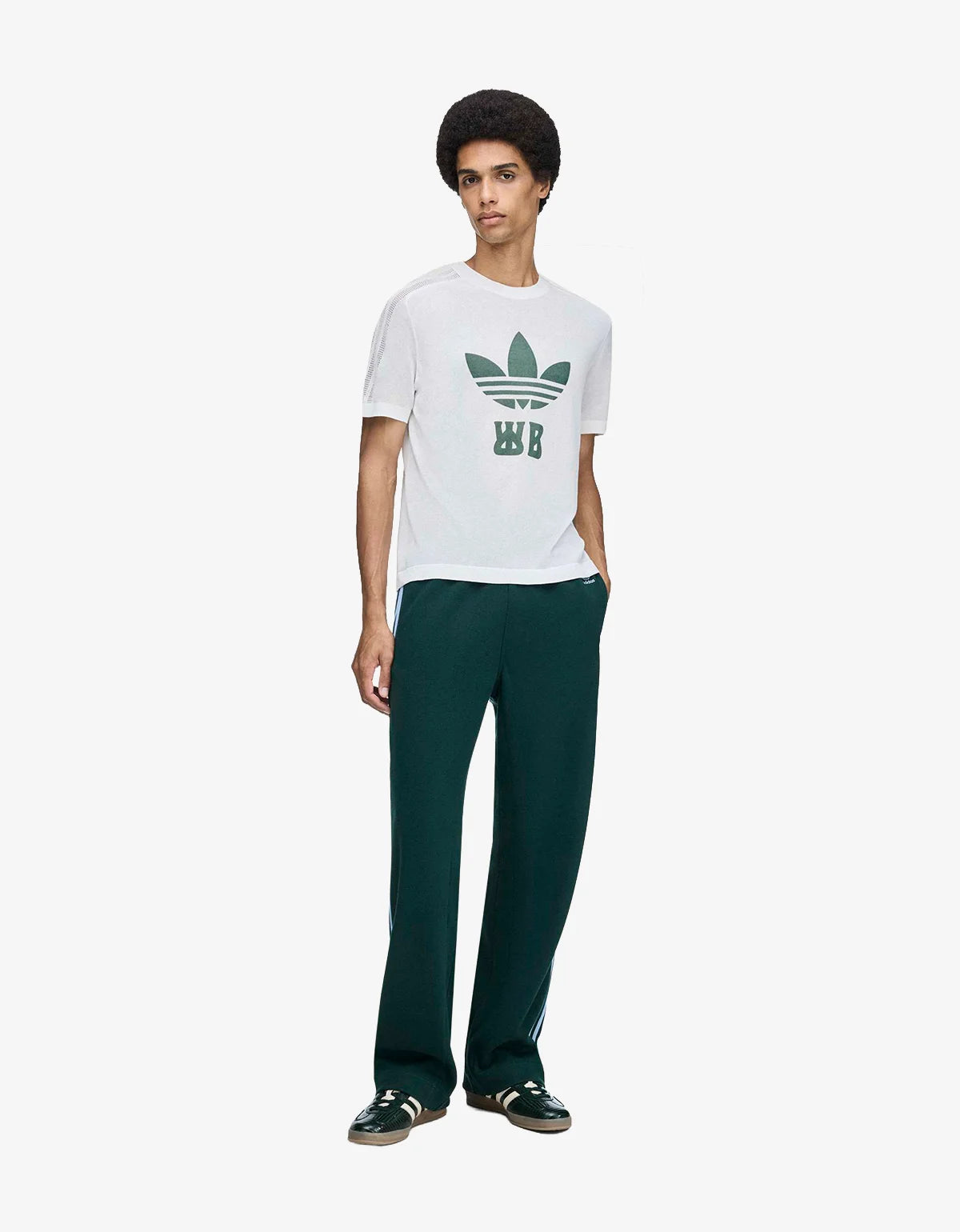A man wearing a white short-sleeve t-shirt with a green Adidas Trefoil logo and 'WB' monogram on the front, paired with dark green pants, against a light gray studio background.