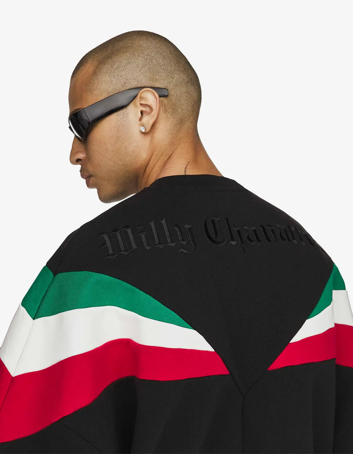 A man shown from the back wearing a black sweatshirt with green, white, and red diagonal stripes across the shoulders and upper back, plus embossed 'Willy Chavarria' lettering on the black upper back. He is also wearing black sunglasses against a plain light grey studio background.