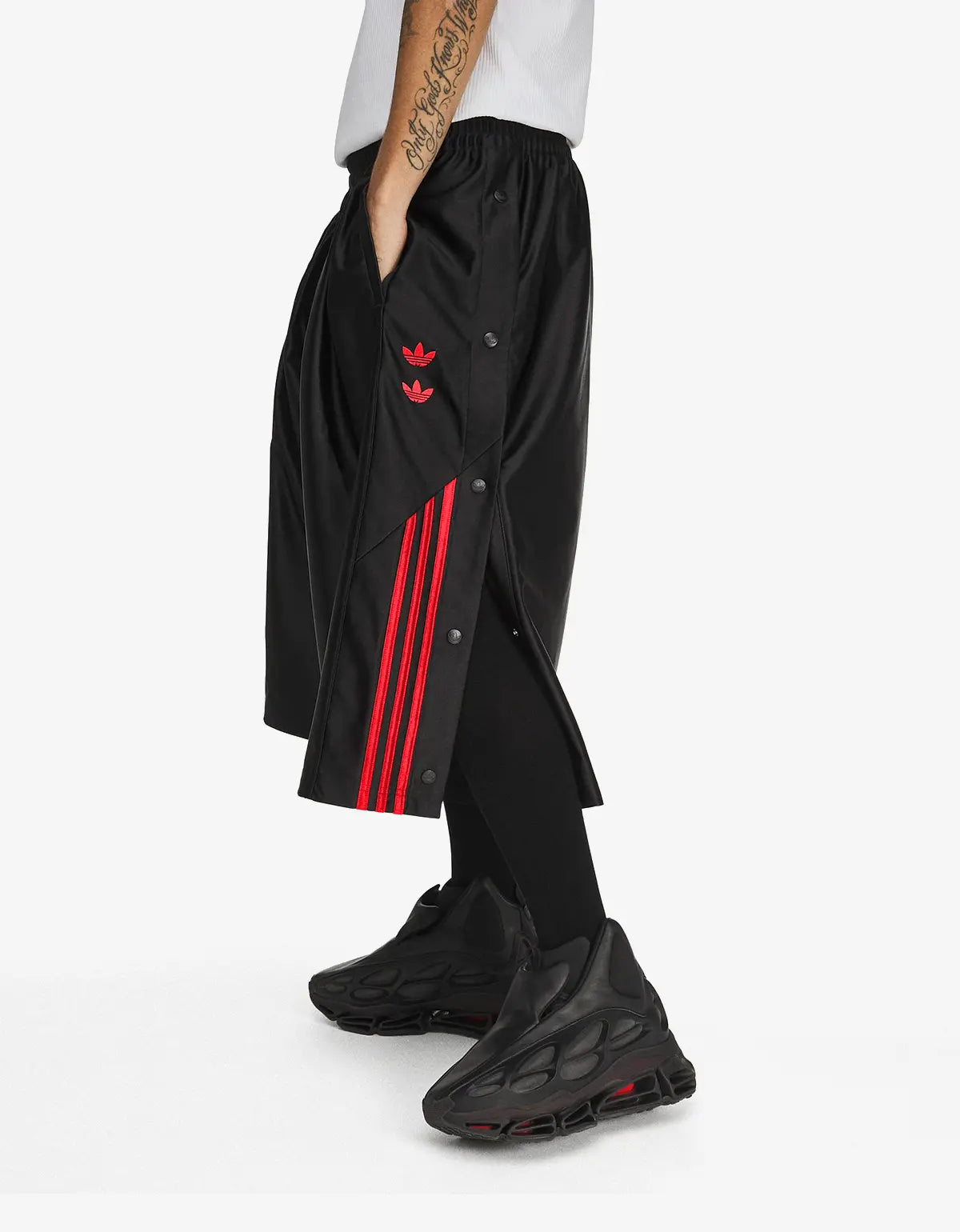 A person wearing black Adidas track pants with red three-stripes and a red trefoil logo, styled with black sneakers against a clean white studio background.