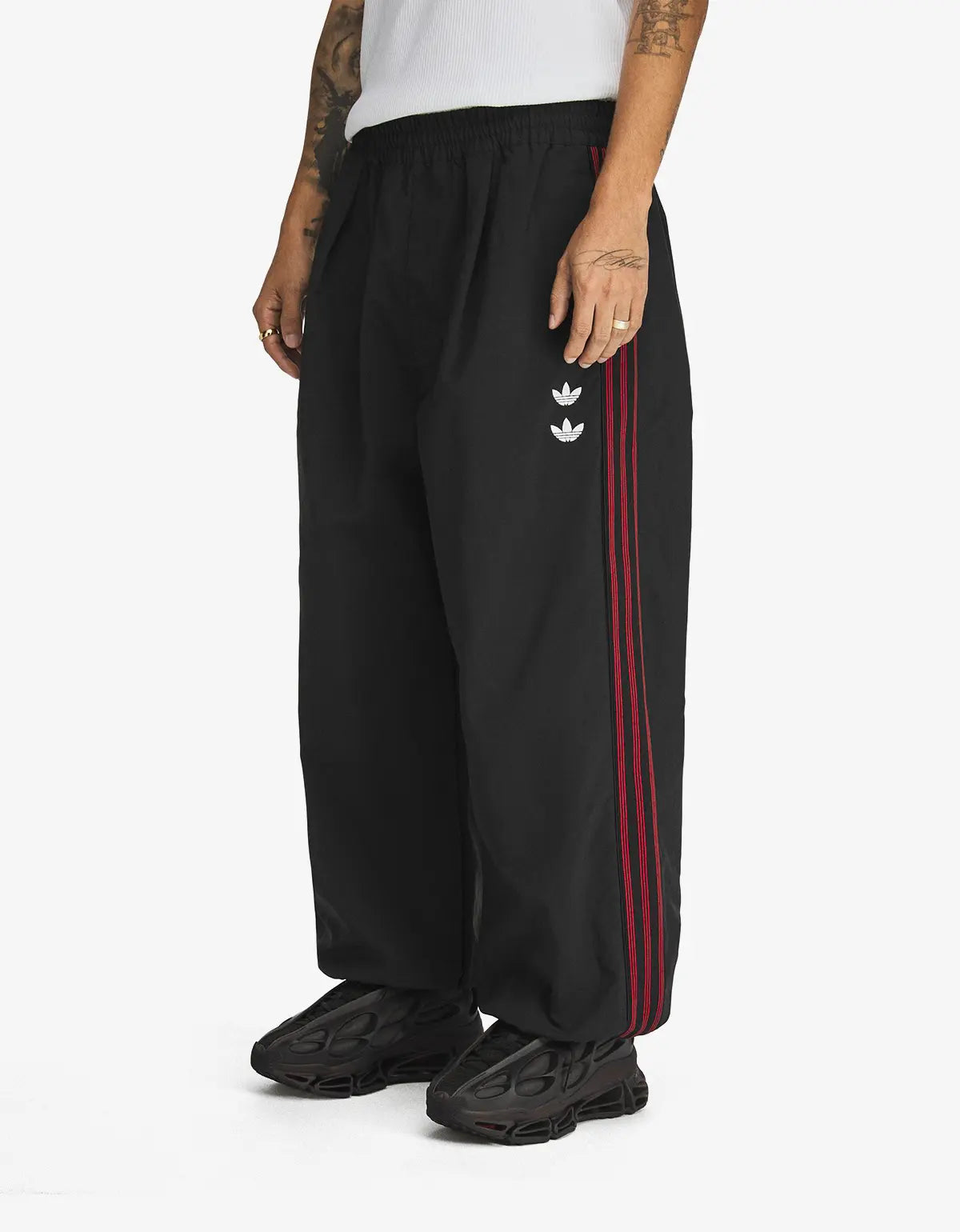 Black wide-leg track pants with red side stripes and a white trefoil logo on the left thigh, shown on a model against a light grey background.