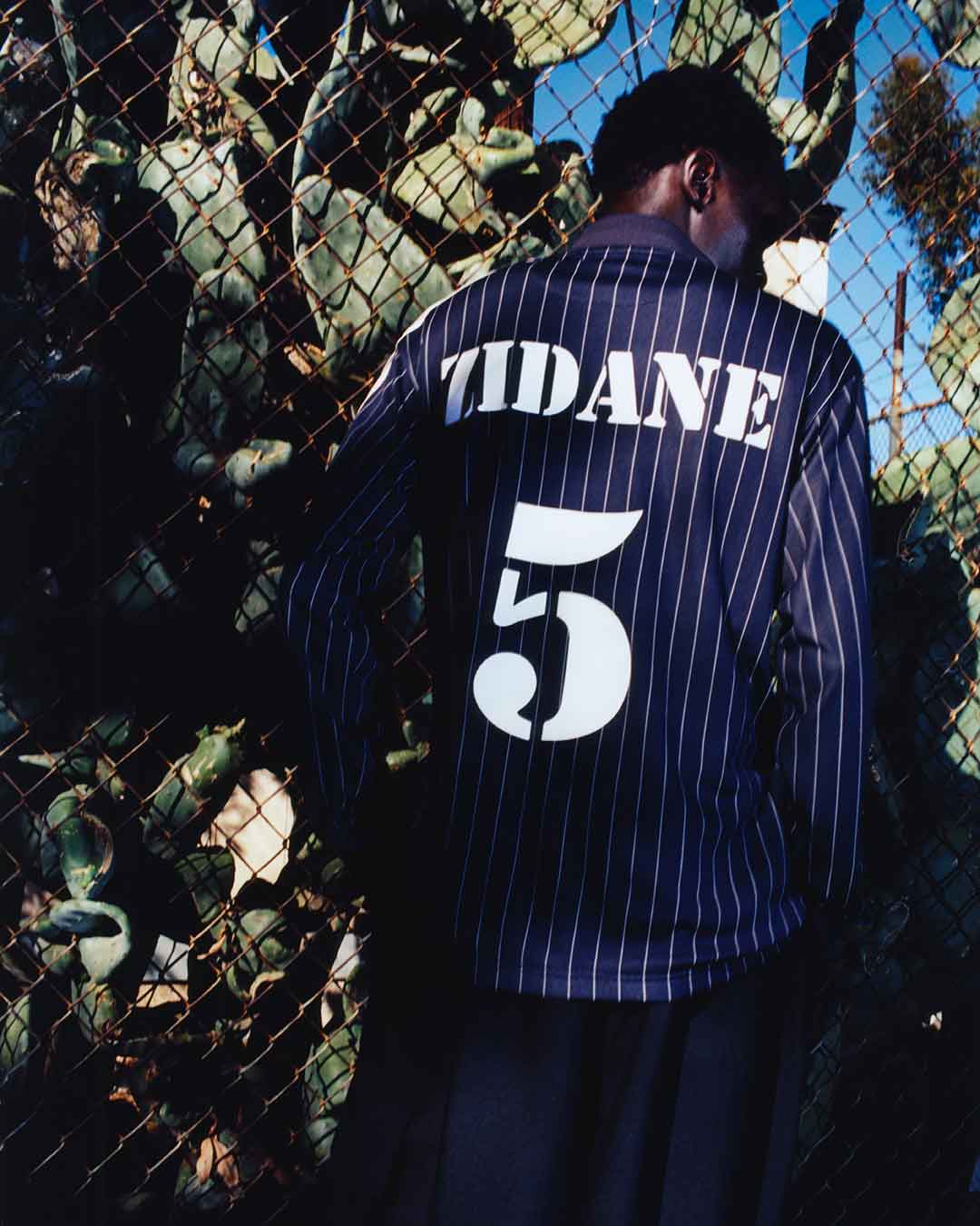 Y-3 Intention SS26 Chapter 2&3. Model wearing a blue pinstripe jersey with 'Zidane' and number '5' standing in front of a chain-link fence.