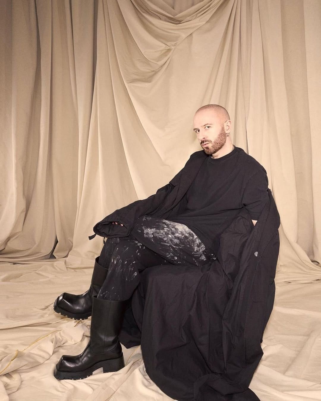 Demna Balenciaga  wearing a black outfit sitting on a draped beige curtain